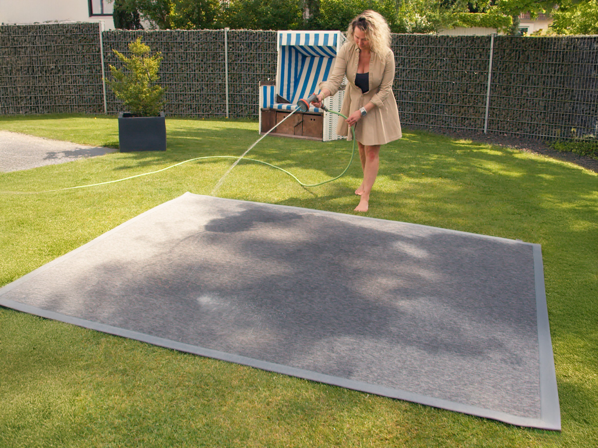 Frau reinigt rechteckigen grauen Outdoorteppich von Ruxedo im Garten mit einem Gartenschlauch – pflegeleichtes, wetterfestes Material
