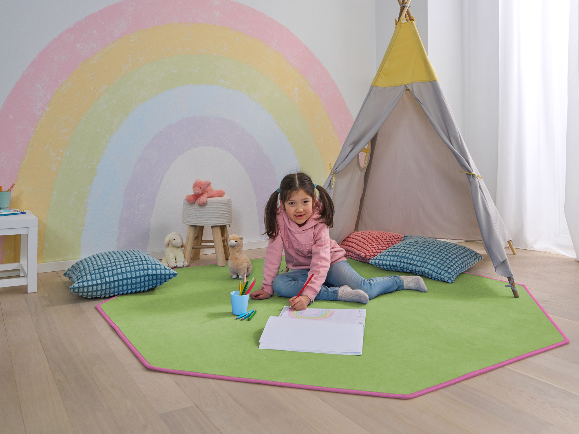 Mädchen malt auf grünem mehreckigen Kinderteppich mit pinker Einfassung vor Tipi-Zelt im Kinderzimmer