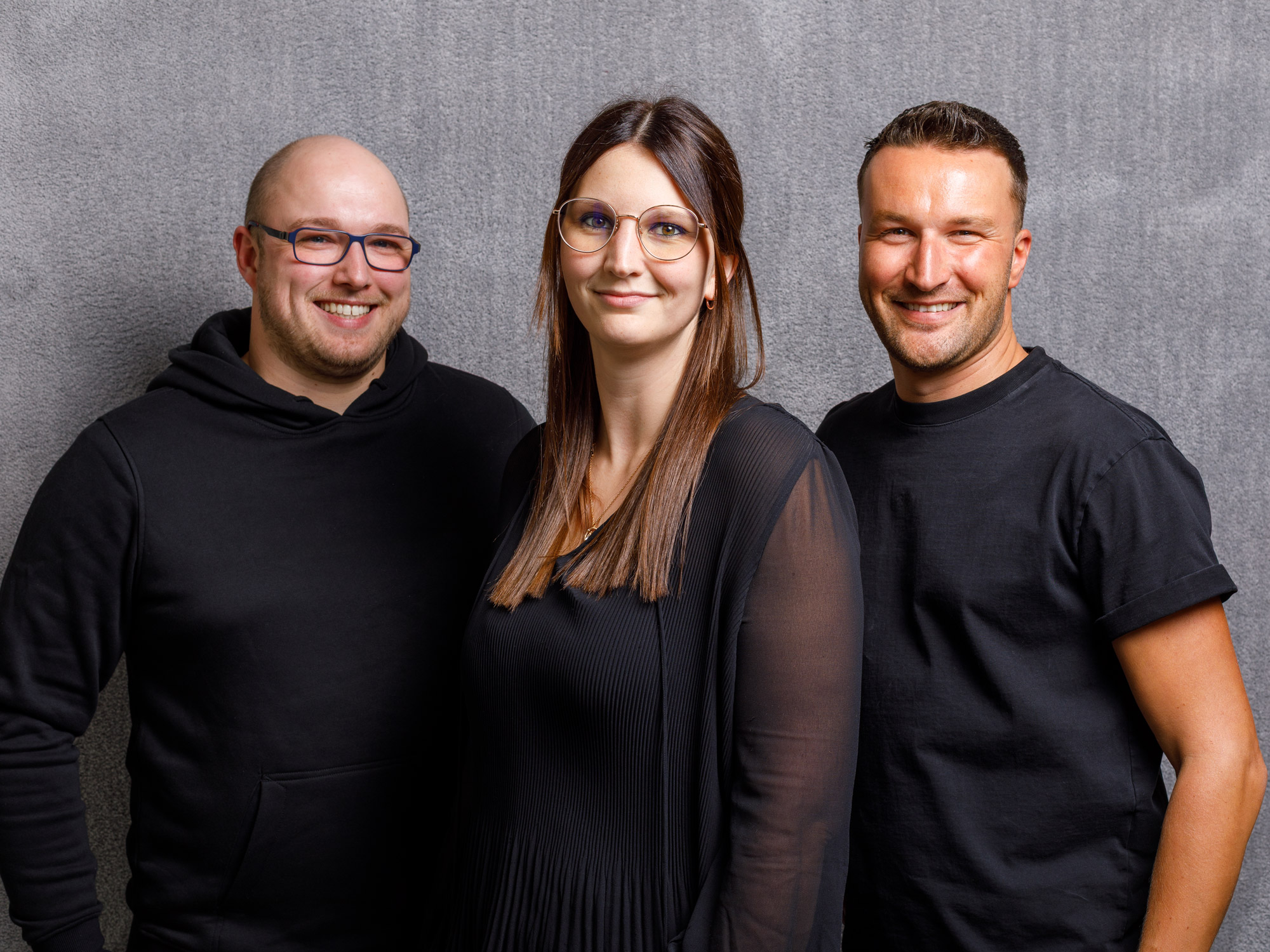 Drei Teammitglieder von Ruxedo lächeln gemeinsam vor einer grauen Wand – Teamfoto aus dem Showroom