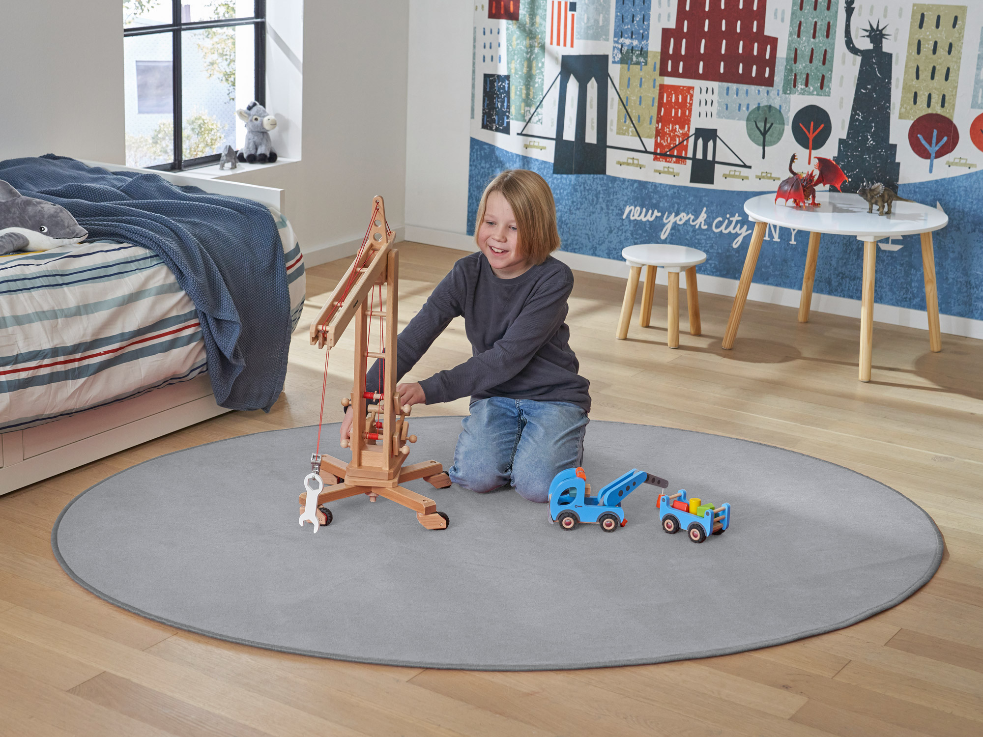 Kind spielt mit Holzspielzeug auf rundem grauen Kinderteppich im hellen Kinderzimmer