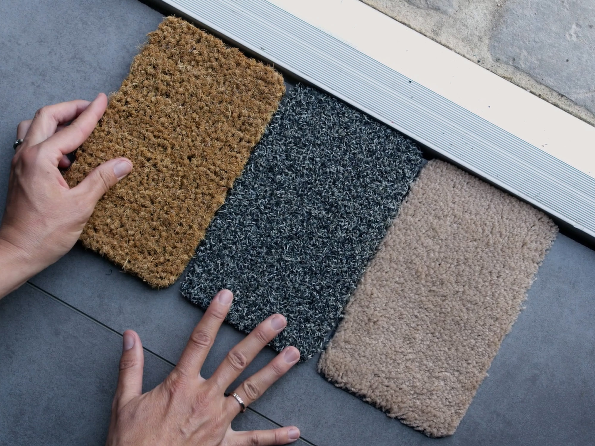 „Drei Ruxedo-Fußmatten-Muster in Braun, Dunkelgrau und Beige liegen vor einer Haustür; Hände halten die Muster zum Vergleichen fest